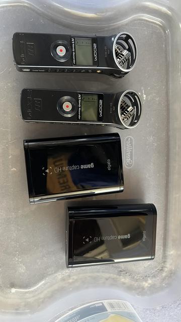 The image shows four digital recorders arranged on a transparent plastic tray. The top two recorders are black with a circular red record button and a small screen displaying recording levels. They are labeled "Zoom H1 Handy Recorder." The bottom two recorders are black with a glossy finish and the text "game capture HD" and "elgato" visible on their screens. The top two recorders have a silver microphone grille, while the bottom two have a sleeker design. The tray has a logo that reads "elgato" and is placed on a gray surface.

Provided by @altbot, generated privately and locally using Ovis2-8B

🌱 Energy used: 1.454 Wh