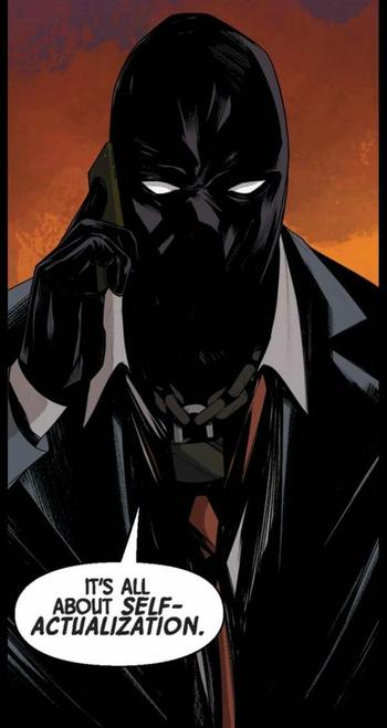 A man in a black mask and black gloves, wearing a dark suit, white shirt and red tie is wearing a padlocked chain around his neck like a necklace. He’s on the cell phone and says, “It’s all about SELF – ACTUALIZATION.“