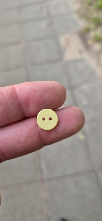Gelber Knopf auf zwei Fingern liegenden. Sieht aus wie Smiley ohne Mund 😶