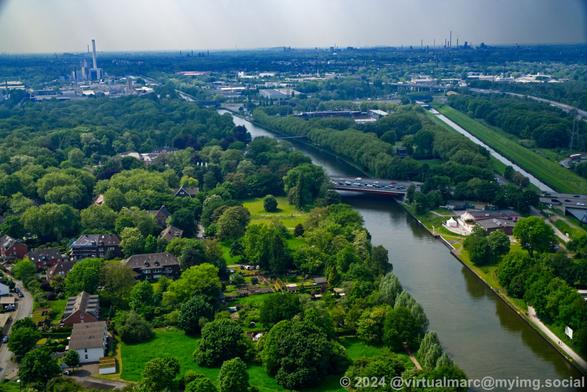A photo from the roof of the Gasometer in Oberhausen. A green landscape with industry in the distance can be seen. There is the Rhein-Herne-Canal and the Emscher river flowing through the image. A freeway is crossing the canal and river over bridges.