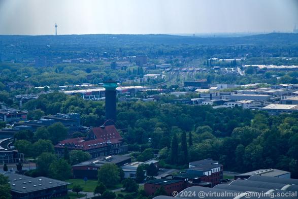 A photo from the roof of the Gasometer in Oberhausen. There are some industry buildings between a green tree landscape. A water tower is standing inside of it. A railway and a trainstation can be seen in the distance. In the far distance you can see a TV Tower on a hill.