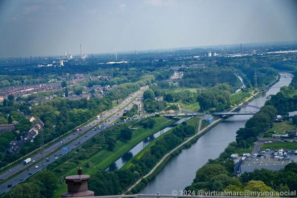 A photo from the roof of the Gasometer in Oberhausen. A freeway is going through the image, parallel to the Emscher river and the Rhein-Herne-Canal. A freeway exit is crossing the river and canal over bridges. Industry can be seen in the back.