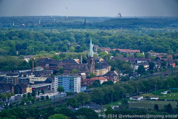 A photo from the roof of the Gasometer in Oberhausen. A church can be seen bettween buildings behind a railway track that is behind a soccer and archery field. Behind many trees and a green landscape you can see the spoil heap Beckstraße with the Tetraeder in Bottrop.