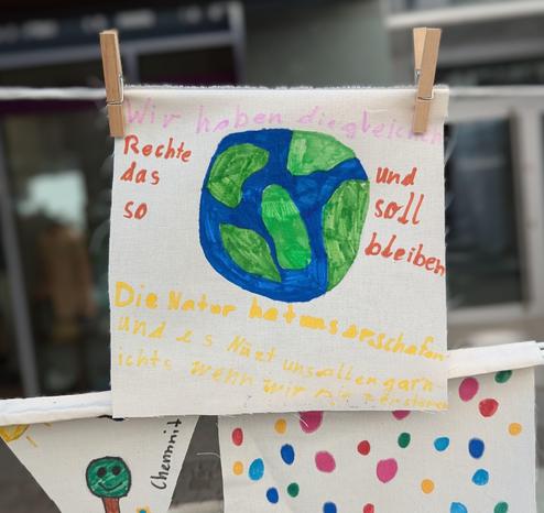 The image features a handmade fabric banner with a drawing of the Earth in the center, colored in blue and green. The Earth is surrounded by text in German. At the top, the text reads "Wir haben die gleichen Rechte das so und soll bleiben" in pink and orange. Below the Earth, the text states "Die Natur hat uns beschaffen und es Nützt nichts wenn uns allen gar nichts wird wenn wir mit unserem Klimawandel" in yellow. The banner is held up by two wooden clothespins. The background is blurred, but it appears to be an outdoor setting with a building visible. The banner is part of a larger display, with other colorful fabric pieces visible at the bottom.
