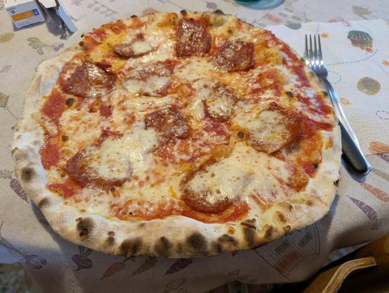 A freshly baked pizza with a thin, crispy crust, topped with melted mozzarella cheese and slices of spicy salami, is served on a table covered with a patterned tablecloth. A fork and knife rest on a napkin beside the pizza.