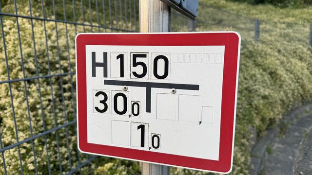 Versorgungsleitung-Hinweistafel in weiß mit schwarzer Schrift und rotem Rahmen: H150 30,0 1,0 Im Hintergrund ein Drahtgitterzaun und gelb blühender Bodendecket.