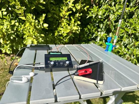 Picture of a QMX radio on a table with a microphone and notepad. An antenna is also attached to the table.