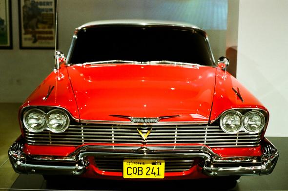 A red classic car with a large silver grill and front embellishments sits in a white museum. Its hood shines bright after a nice wax.

Source text: https://www.petersen.org/vehicle-spotlights/1958-plymouth-fury-christine