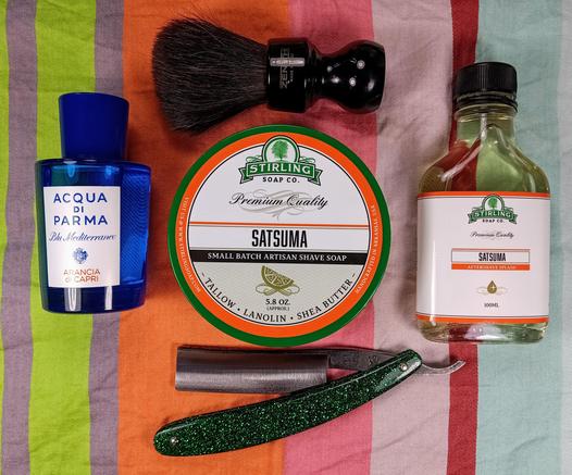 (Le Chat Mistral description)

The image showcases a collection of grooming items arranged on a striped background. At the top left, there's a blue bottle of Acqua di Parma cologne. To the right of the cologne, there's a shaving brush with a black handle and a tin of Stirling Soap Co. Satsuma shaving soap. Below the shaving soap, there's a green straight razor with a metallic handle. To the right of the razor, there's a small bottle of Stirling Soap Co. Satsuma aftershave splash. The items are neatly laid out, suggesting a traditional shaving setup.