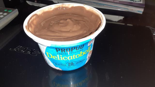 Offener Puddingbecher schräg von oben rein fotografiert.  Der offene Becher steht auf einem geschlossenen Notebook auf einem dunkelbraunen Couchtisch.