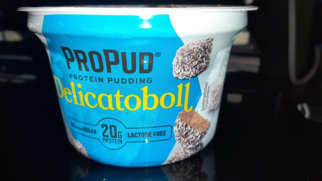 Puddingbecher von Vorne mit Etikett fotografiert wo auf blauen Hintergrund der Name PROPUD und Delicatoboll steht. Rechts neben dem Text sind 3 "Schokoküsse" abgebildet. Die Schrift ist in schwarz & gelb auf dem blauen Hintergrund.