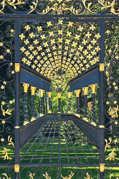 A close-up, front-on view of an elaborate black metal gate with intricate gold detailing, including many small four-leaf clover or cross shapes.

The gate is open, revealing a lush green lawn and trees in the background, suggesting a garden or estate. The gold elements on the gate appear to radiate outwards from the centre.