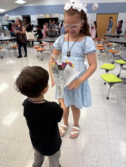 Young girl in a light blue dress with a white bow and glasses joyfully holding a bouquet of flowers given by a young boy in a black shirt. They stand on a tiled floor in a cafeteria with tables and chairs; several other people are in the background, engaged in different activities.