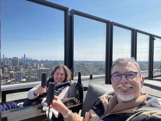 A man and a woman are seated outdoors on a rooftop with a city skyline in the background. They are smiling and holding up drinks in sleek black glasses. A bottle of wine is visible nearby, and the scene is bathed in bright sunlight with clear skies