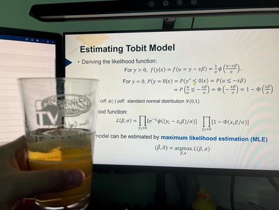 A person holds a glass of sweet tea vodka &amp; lemonade in front of computer monitors displaying a presentation titled "Estimating Tobit Model." The screen features mathematical formulas and likelihood functions related to the Tobit model.