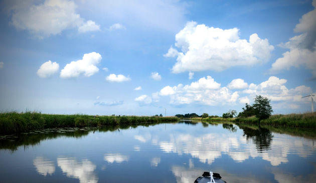 Inmitten von Wiesen und Feldern spiegelt sich der blaue Himmel mit Schäfchenwolken auf einer glatten Wasseroberfläche.