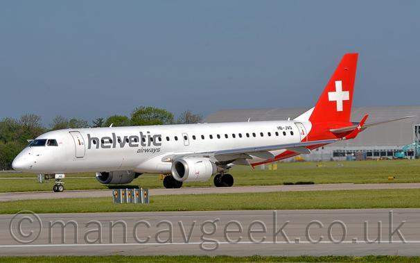 Side view of a twin engined jet airliner taxiing from right to left on a taxiway.
The plane is mostly white, with dark grey "Helvetic Airways" titles on the forward fuselage, with a red rear fuselage and tail overlaid with a large white cross, a logo repeated on the up-turned wing-tips.
The black registration "HB-JVO" is on the upper rear fuselage.
A grey runway runs along the bottom of the screen, separated from this taxiway by grass, with more grass in the background leading up to a large grey building on the right, and trees on the left, under blue-grey skies.
