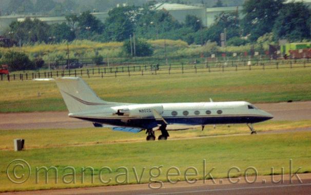 Slightly blurry and grainy, side view of a twin engined BizJet taxiing from left to right.
The plane is mostly white, with a dark blue belly, and a yellow stripe running along the body, with another yellow stripe on the tail.
A black runway bordered by large areas of grass fills the foreground, with more grass leading up to trees in the background.