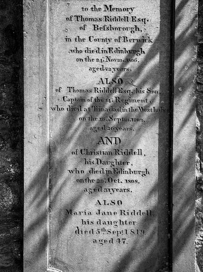 Aged stone memorial plaque with engraved text commemorating Thomas Riddell Esq. and his family members who passed away in Edinburgh in the 19th century. The text, worn but legible, details names, dates of death, and ages. The plaque is split down the middle with shadows to the right side, highlighting the weathered surface and historical wear.