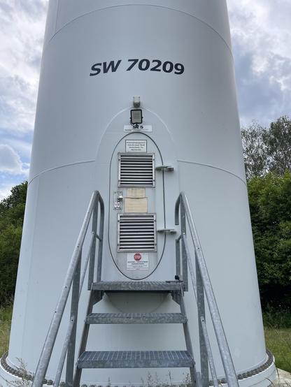 The door at the basement of a large wind turbine. Small metal staircase leading up to the door. Several plates with warning signs and specs can be seen at the door.