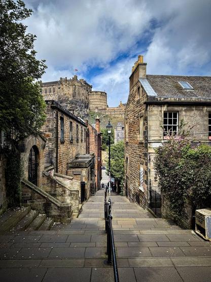 Narrow cobblestone street leading up to a grand, ancient stone castle with flags flying on its battlements, flanked by traditional stone buildings with a blue sky and scattered clouds above. A metal railing runs along the left side of the street, guiding the path of pedestrians walking along the pavement.