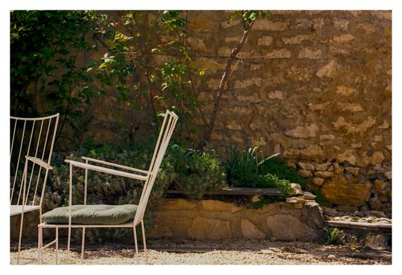 Color photograph of a brick wall in front of which ornamental roses are sprouting and two white metal chairs with green cushions are standing.