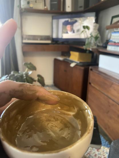 A hand holding a small, ceramic bowl with a brown residue of gold and honey is in focus, while a television in the background shows a scene from Amadeus featuring a man in formal attire. The setting includes wooden furniture and indoor plants.