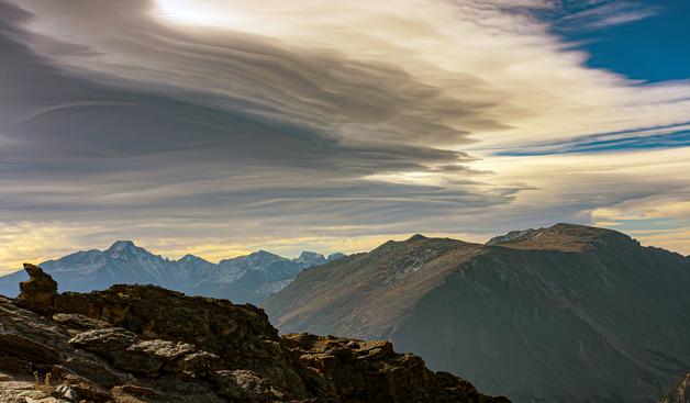 Scenic photo in Rocky Mountain National Park, looking south from Trail Ridge Road. Sharply focused rocks in the foreground with sunlight glinting off surfaces and shadows marking the edge. Behind ridge in front is the Front Range of the American Rocky Mountains. Lings Peak is the highest one at the left. The light is subdued, with lots of shadow hiding details. Snow covers most of the mountainsides farthest away. Lenticular clouds shading from dark gray to bright white show striations caused by wind shear. Deep blue sky shows in the upper right corner. October 2012