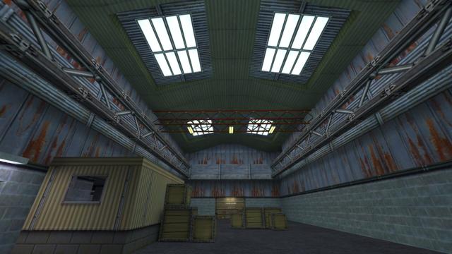 In this picture you can see the inside of the main building. It is a large hall. From the left navh right there is a bright yellow corrugated iron building, to the right there are small wooden boxes stacked on top of each other and a little further to the right there are also wooden boxes. In the background you can see a heavy metal gate. In addition, you can see that there are steel girders under the roof and four windows are installed in the roof. This is on the map de_nuke in the game Counter-Strike 1.6.