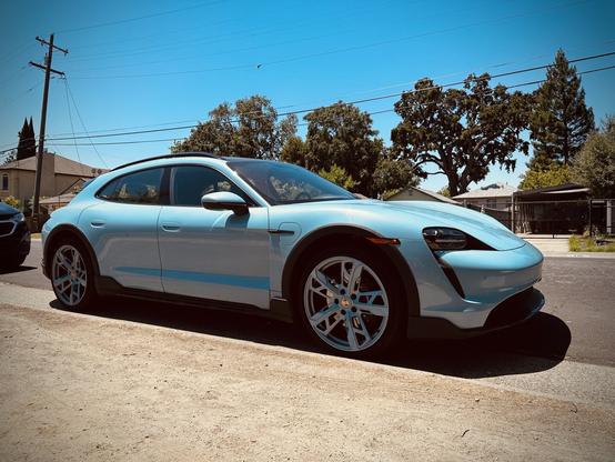 A light blue electric wagon is parked on a residential street. The vehicle features distinct wheels in matching color and sleek design elements. Surrounding the car are trees and houses, with clear blue skies above.