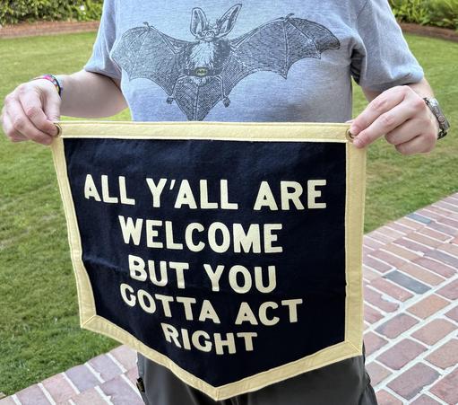 person holding up a small square felt banner, with a yellow outline and a blue background containing yellow all-caps text that says ALL Y'ALL ARE  / WELCOME / BUT YOU / GOTTA ACT / RIGHT in the center.
