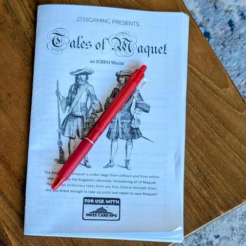 A wooden table with a zine style RPG entitled "Tales of Maquet an ICRPG world" and a red pen on top.