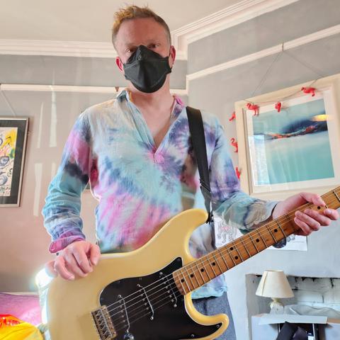 Man wearing black CAN99 mask, tie-dye shirt and holding Fender Stratocaster guitar.