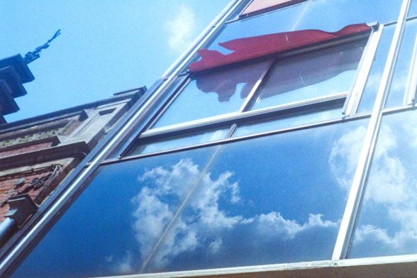 Upward view of a modern glass building reflecting a bright blue sky with scattered clouds, alongside part of an older brick structure. A red awning casts a soft shadow, adding contrast to the reflective surfaces. Shot on 35mm film.