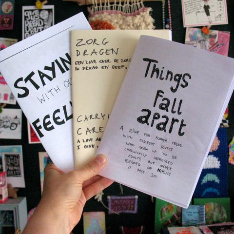 Photo of hand that holds 3 zines in the air. Titles include "Zorg dragen" and "Things fall apart".