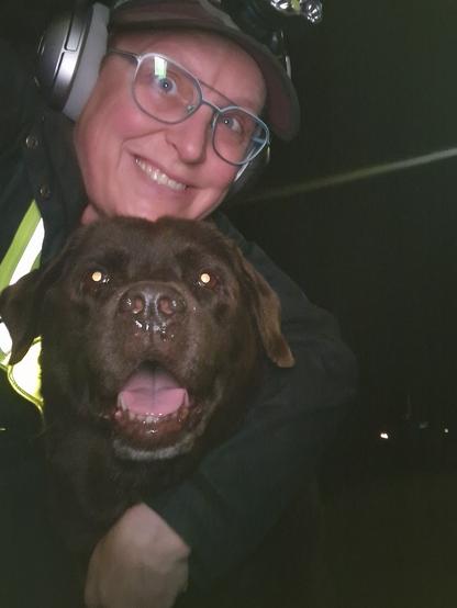 Night time selfie of a person and a chocolate brown Labrador with greying snout. The person is wearing a black jacket, yellow safety harness, red cap, silver headphones and blue-rimmed glasses. On their cap rests a headstrap torch in off position. Their free hand is around the dog's shoulders. Both are facing the camera. The dog's mouth is slightly open, showing off some blep. The person's head is resting on the dog's head. They're smiling, grateful for their time spent together.
