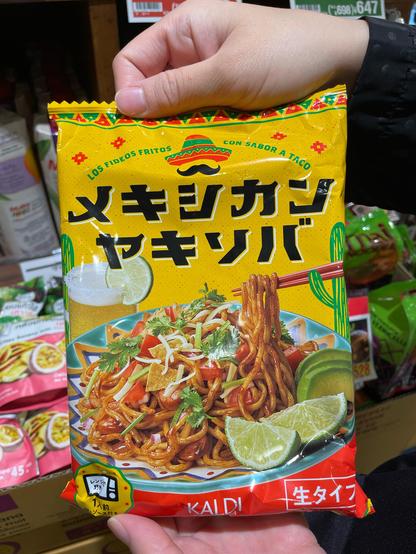 Two hands holding up a package of "Mexican" yakisoba in an import food store. The package is mostly yellow and "Mexican" themed with cartoon cacti and "Mexican Yakisoba" written in Japanese. The product image is, unsurprisingly, yakisoba on a "Mexican" themed plate, topped with tiny nacho chips, tomato, cheese, avocado and lime.