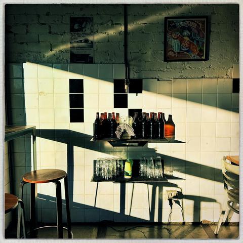 Morning shadows fall across a white-tiled wall, with a shelf of brown-glass water bottles and another shelf below containing glasses. Two pictures are up above, in the shadows, and two stools are visible to the left.