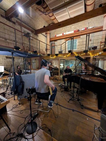 Recording shenanigans under way in The Wood Room at Peter Gabriel's Real World Studios near Bath. 

The band are seated in a large recording studio with a mezzanine floor, with Dan behind a drum kit on the left and John behind a Bosendorfer grand piano on the right. Jim is seated in the foreground with his back to the camera and Adrian (with Fender Jazz master guitar) is at the back.
