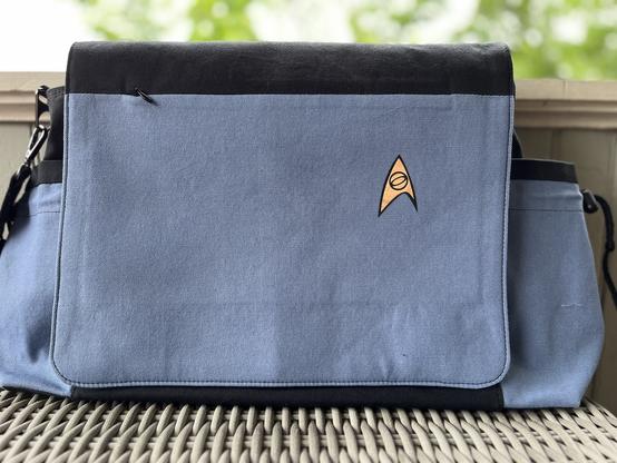 A photograph of a pale blue messenger bag with the Starfleet sciences division badge in the top left corner, similar to the TOS uniform.