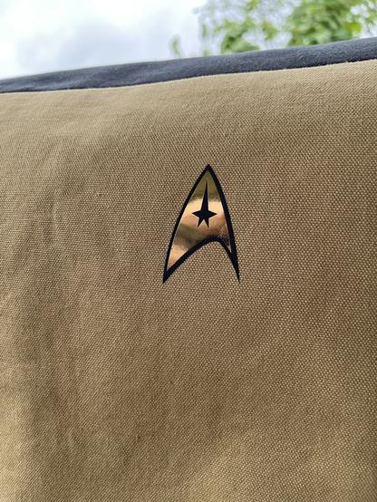 A photograph of a gold messenger bag flap with the Starfleet command division badge in the top left corner, similar to the TOS uniform.