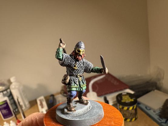 A 28mm scale Viking warrior, wearing a Gjermundbu style helmet and chain mail, and wielding an axe in one hand (over his shoulder) and a sceax in the other. He’s got a green tunic under the mail, blue trousers, brown boots and a red beard. There’s a Thor’s hammer picked out in gold around his neck.