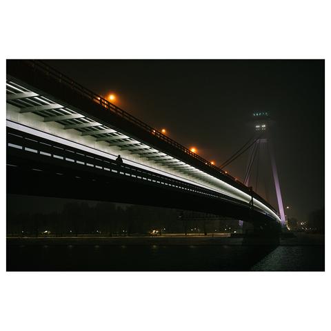 A night photograph of the UFO Bridge in Bratislava, Slovakia. The bridge is lit from below with a row of orange streetlights above and a pedestrian silhouette crossing underneath. The UFO-shaped observation tower is faintly visible through fog in the background.