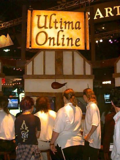 The Ultima Online booth at E3 1996. It shows a faux-medieval side of a house as the booth, and a computer with nerds huddled around someone playing the pre-alpha.