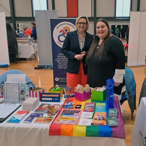 Two women are standing at an information booth. The woman on the left, Jo Briskey, is light-skinned with short, blonde hair and is wearing a dark blazer, light-colored shirt, and bright red pants. The woman on the right, Shannon, is also light-skinned/medium-toned, with long dark brown hair and is wearing a black top with white trim.
The booth is a table covered with a rainbow flag and various items. These include pamphlets, brochures, sunscreen, blue water bottles, pens, and green stress balls. 
A blue banner behind them reads "Brimbank Disability Advocacy & Support" and further information about support is written on the banner and also presented on the table.
The background is indicative of the venue, the Keilor Basketball & Netball stadium. It has fluorescent lighting, polished wooden floors, indoor bench setting, and there are banners and tables throughout.