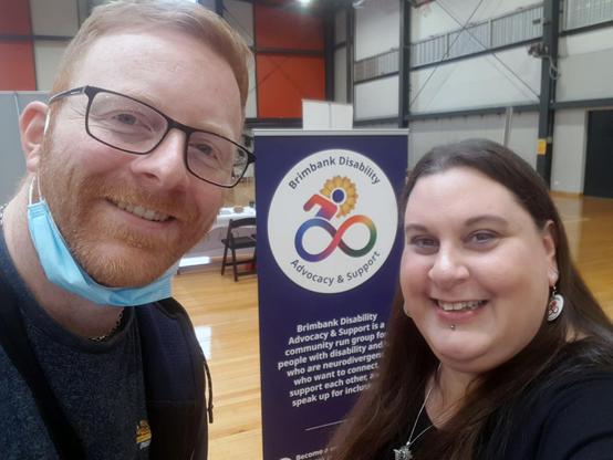 Two people, a man and a woman, are smiling and looking at the camera in a selfie style. 
The man, David P, has reddish-brown hair, glasses, and a light blue face mask sitting below his chin. He is wearing a dark-coloured, casual shirt. Shannon is on the right.