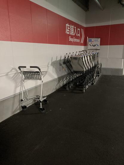An empty shopping cart is positioned to the left, while several more shopping carts are neatly stacked against a wall with a red and white design. A sign indicating the shop entrance is visible nearby. The flooring is dark and the setting appears to be inside a car park of building.