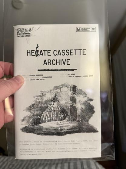 A hand holds a printed document titled "HEGATE CASSETTE ARCHIVE" within a plastic sleeve. The document features text regarding its authors and contributors, along with an illustration of a large dome-like structure surrounded by clouds.