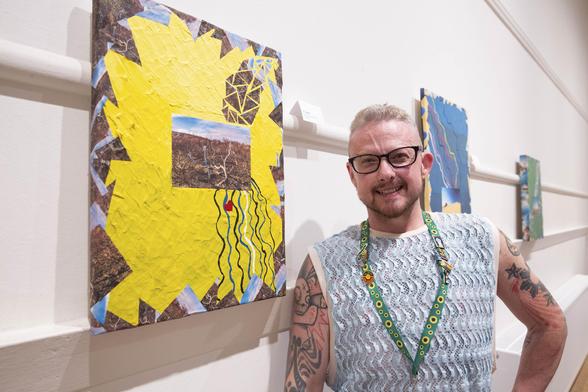 A white trans man in glasses smiles at the camera. On the wall to his right are three canvases of different colours.