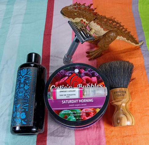 (Le Chat Mistral description)

The image showcases a collection of grooming items arranged on a striped, multicoloured fabric. On the left, there's a dark bottle of Don Jose aftershave. In the centre, a tin of Catie's Bubbles shaving soap, labelled "Saturday Morning," is accompanied by a small bottle of Sweet Stuff Eau de Toilette. To the right of the tin, there's a wooden-handled shaving brush with black bristles. Above these items, a safety razor and a toy dinosaur are placed. The scene is colourful and neatly arranged, giving a sense of a well-organised grooming setup.